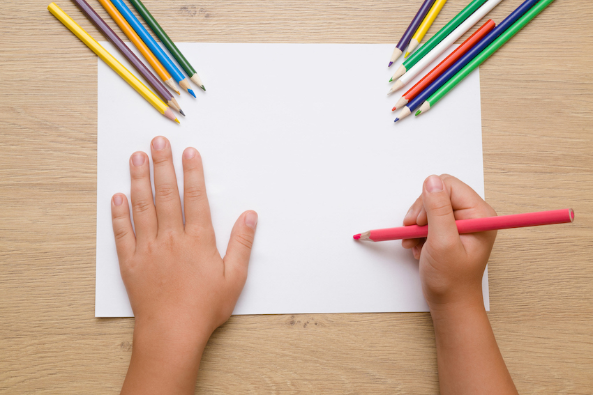 Little girl's hand painting on the white paper with pink pencil. Color pencils on the wooden desk. Drawing time. Top view. Empty place.