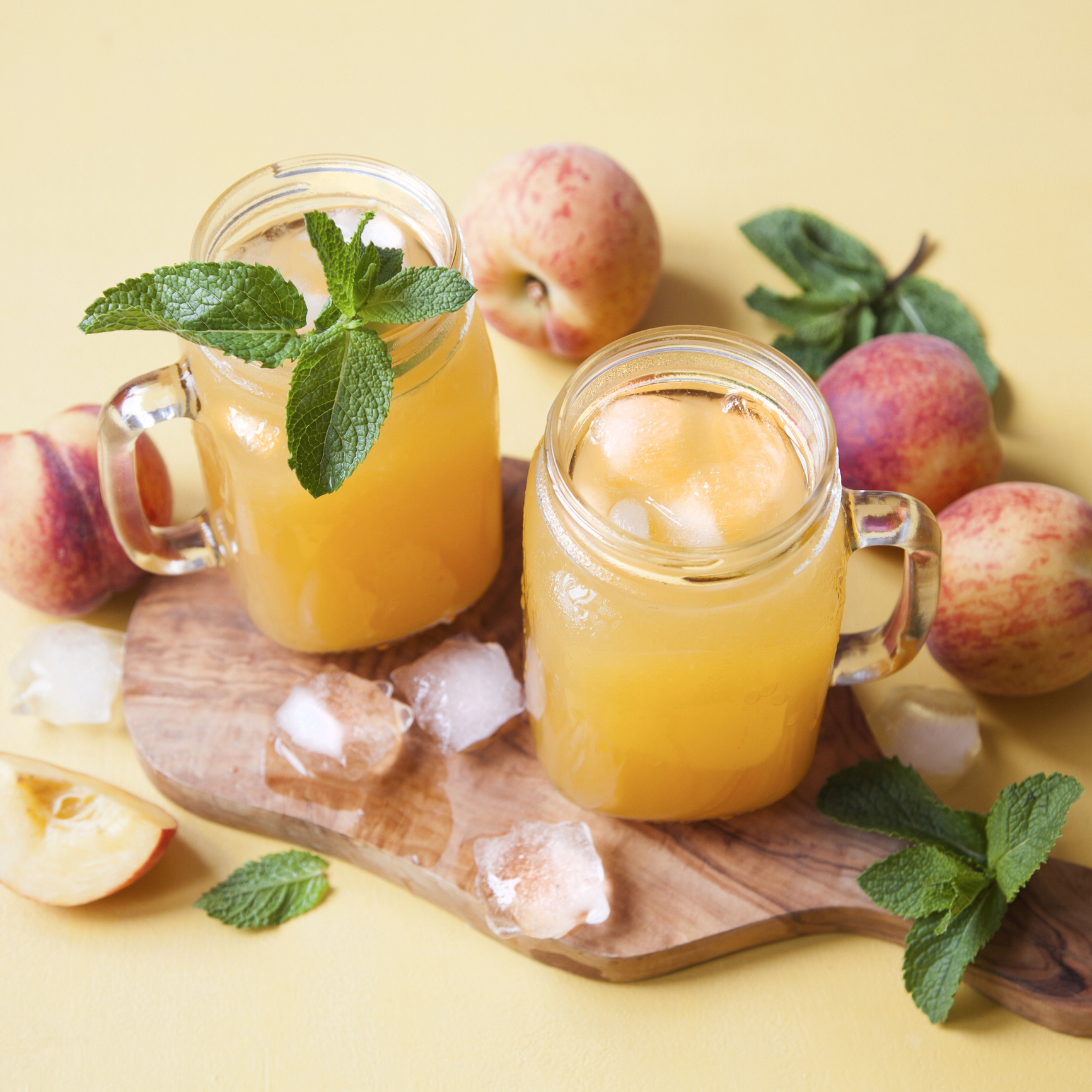 Cold summer peach drink with ice and mint in mason jars on a tray with fresh peaches on yellow background.