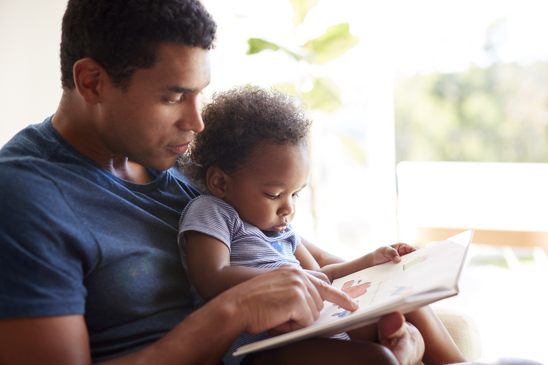 Close up of young adult black father reading a book with his two year old son, close up, side view, backlit