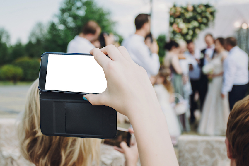 Hand holding phone with empty screen and taking photo of gorgeous bride and stylish groom with guests at wedding reception in restaurant. Photo booth.