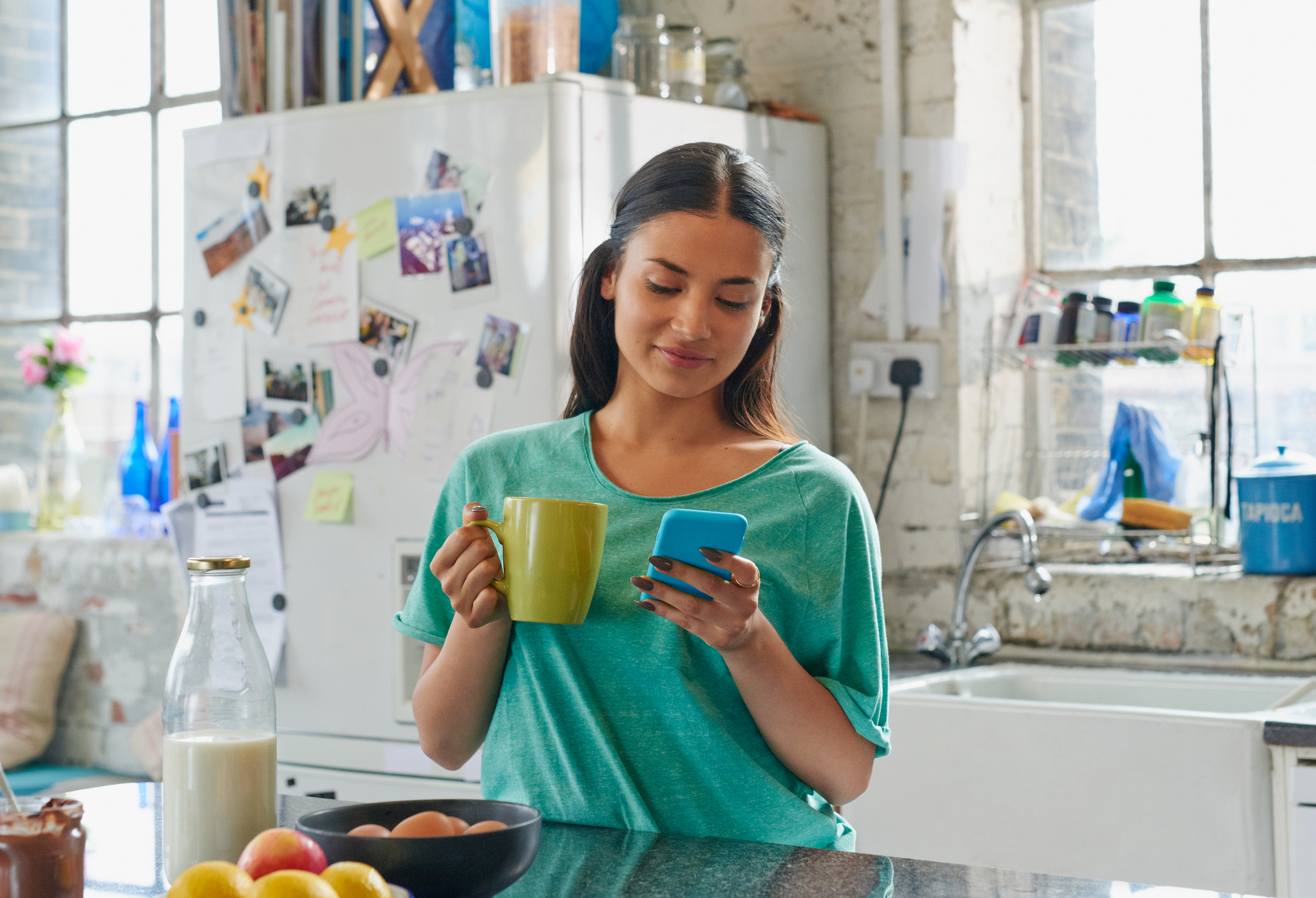 Hispanic girl holding coffee using blue mobile phone at home