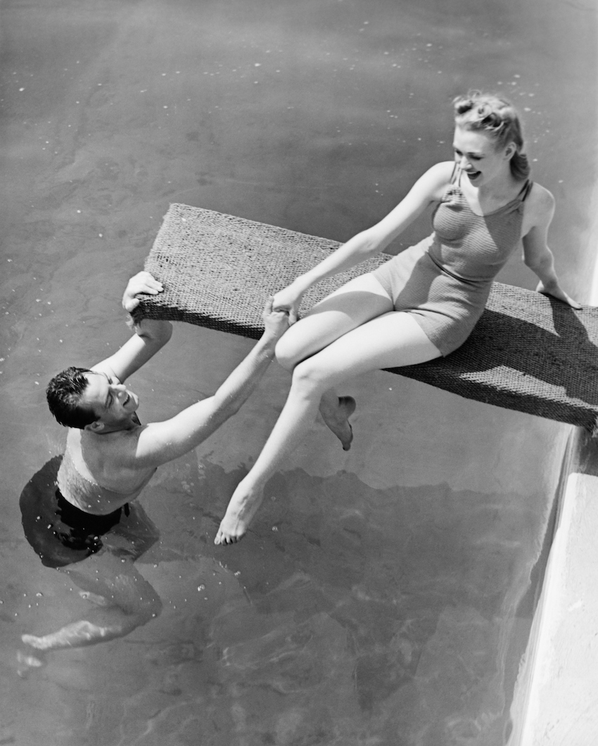 Woman sitting on diving board, man grasping her hand (B&W), elevated view