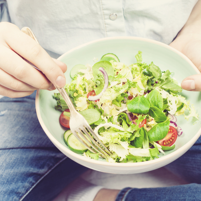Fresh summer salad in bowl with lettuce, tomatoes, onions and cucumbers. Girl in jeans and blue shirt holding fork, top view. Clean eating, dieting food concept