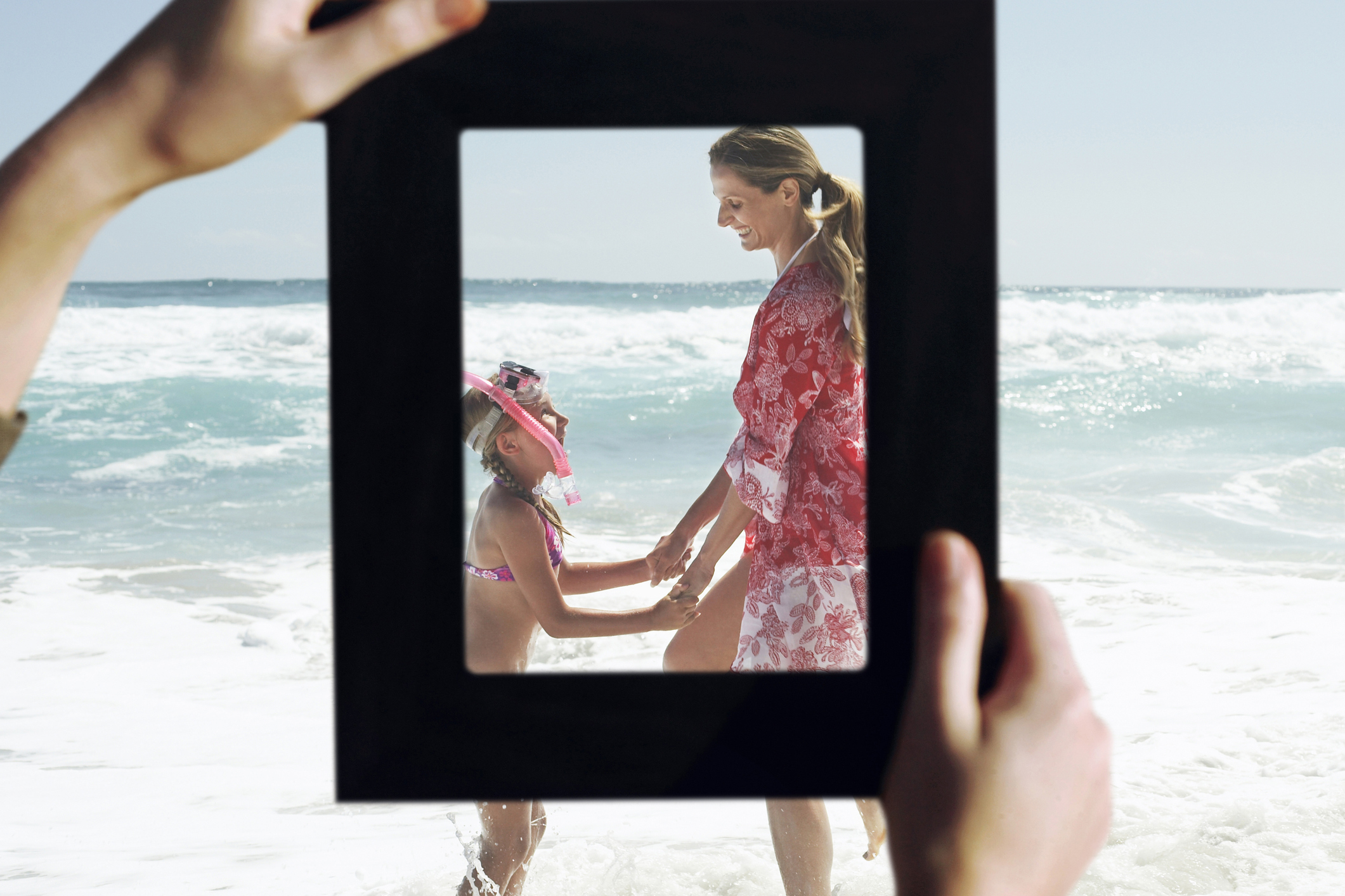 Female hands framing mother and daughter playing in water at beach