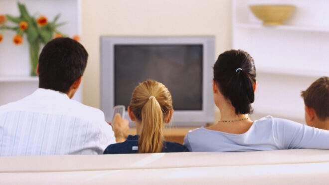 Rear view of a couple sitting on the couch in front of a television with their children (8-10)