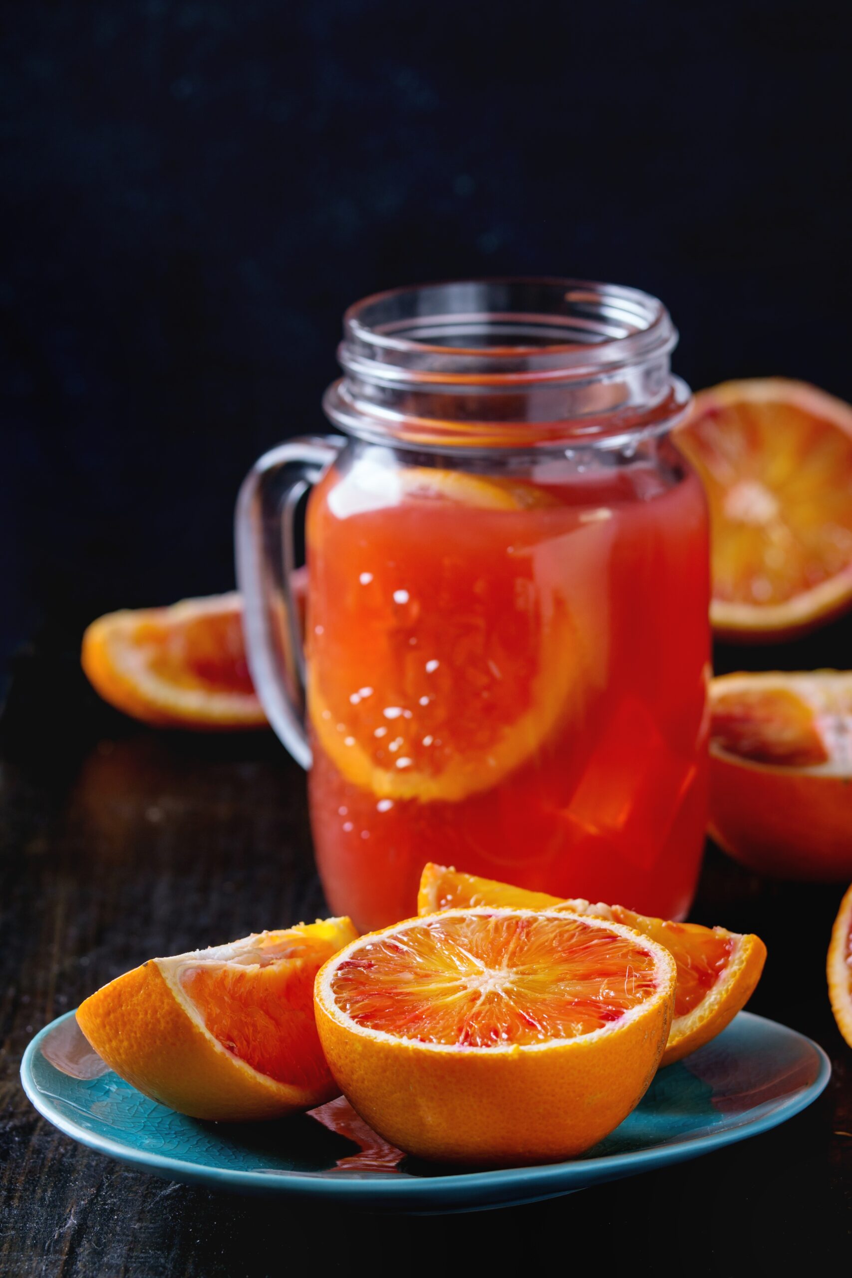 Sliced Sicilian Blood oranges in turquoise ceramic plate and glass mason jar of fresh red orange juice over old wooden table. Dark rustic style.