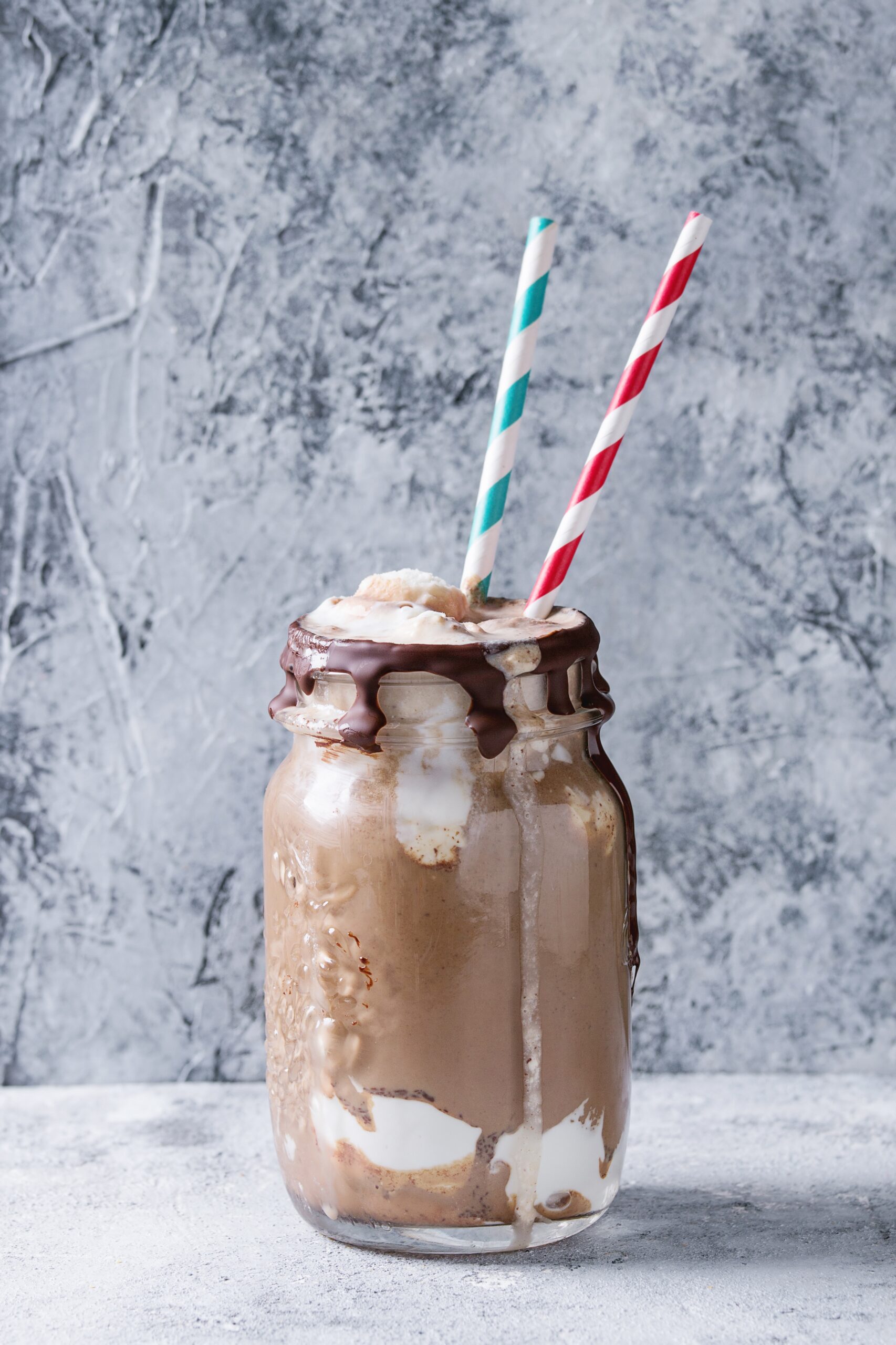 Chocolate coffee milkshake with ice cream scoop served in glass mason jar with retro cocktail tubes on gray texture background. Summer sweet drink