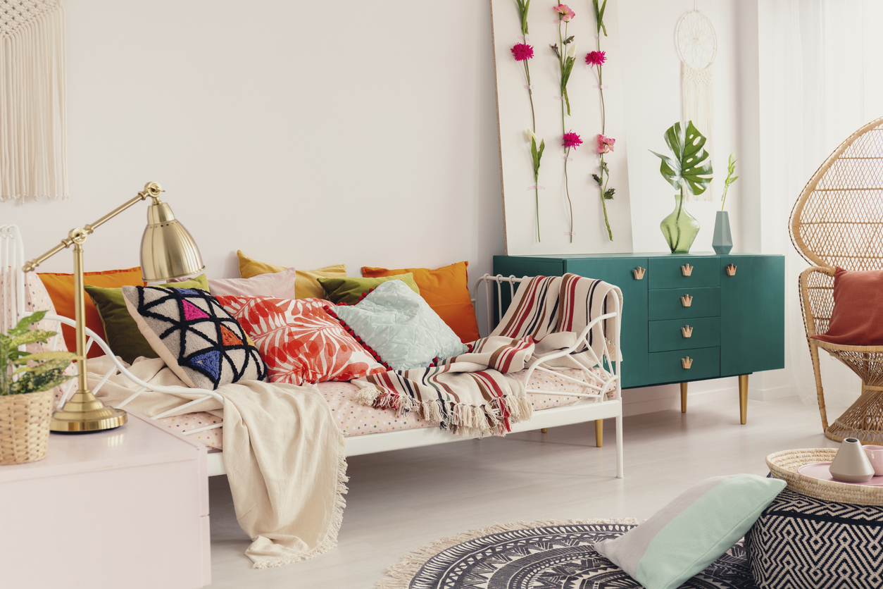 Patterned and colorful pillows on single metal bed in stylish girl's bedroom interior with peacock chair and green cabinet with crown shape handles