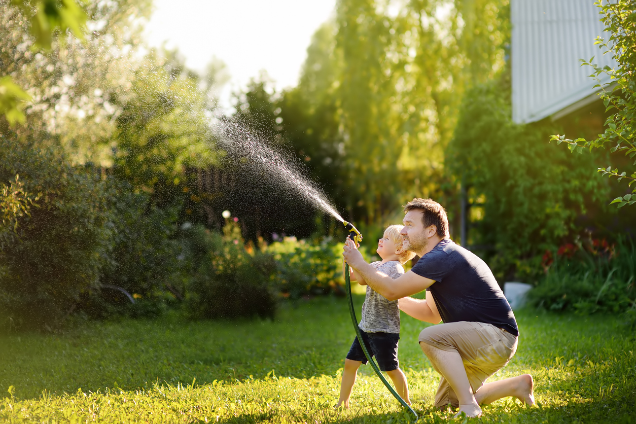 Funny little boy with his father playing with garden hose in sunny backyard