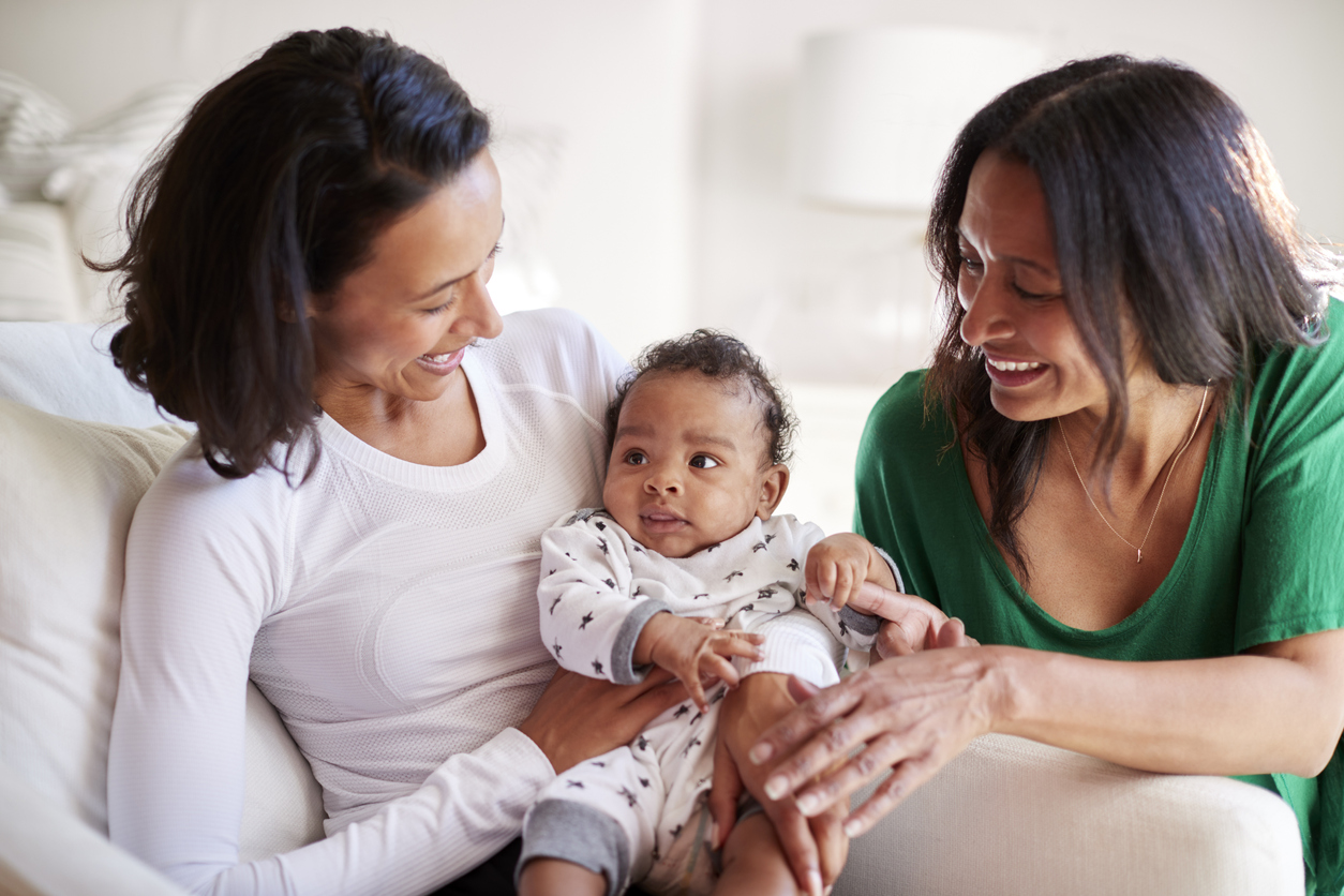 Millennial mixed race mother sitting in an armchair holding her three month old baby son, her mother kneeling beside them