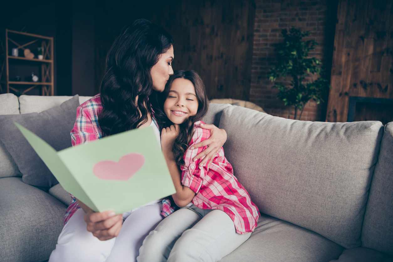 Close up photo two people mum little daughter giving postcard with mommy poem kissing sweet girl unexpected cute delighted wear pink plaid shirts flat apartment room sit cozy couch sofa divan