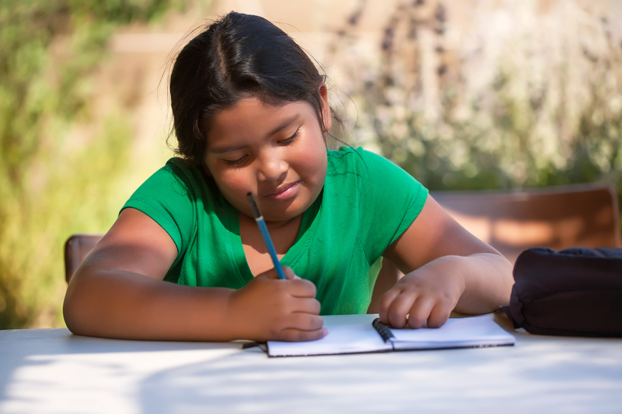 Smart elementary student using imagination to sketch ideas on notebook in an outdoor setting while the sun is setting.