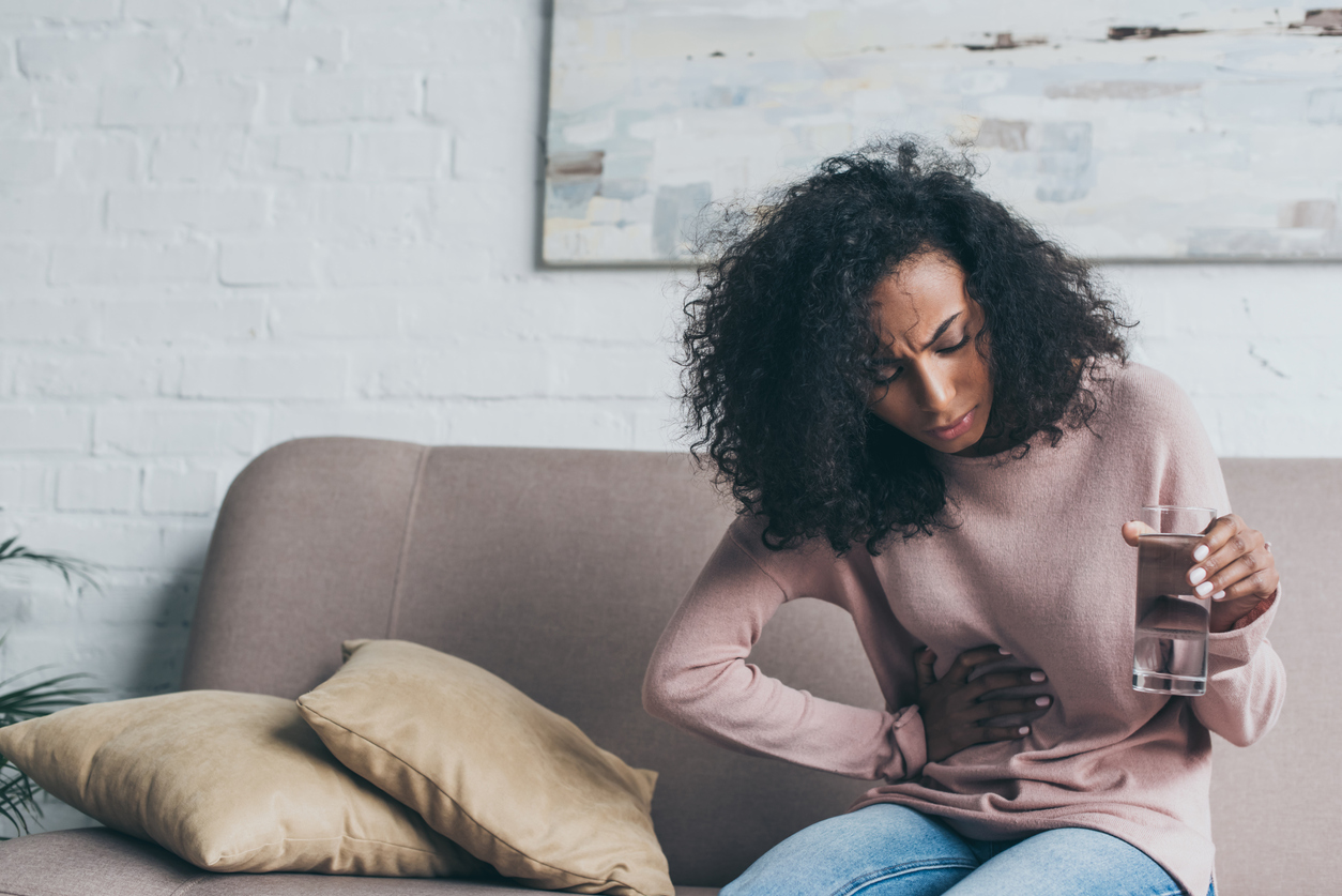 unhappy african american woman holding glass of water while sitting on sofa and suffering from stomach pain