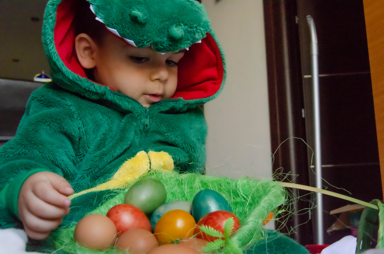 Cute toddler in the green dinosaur costume playing with the colorful easter eggs.