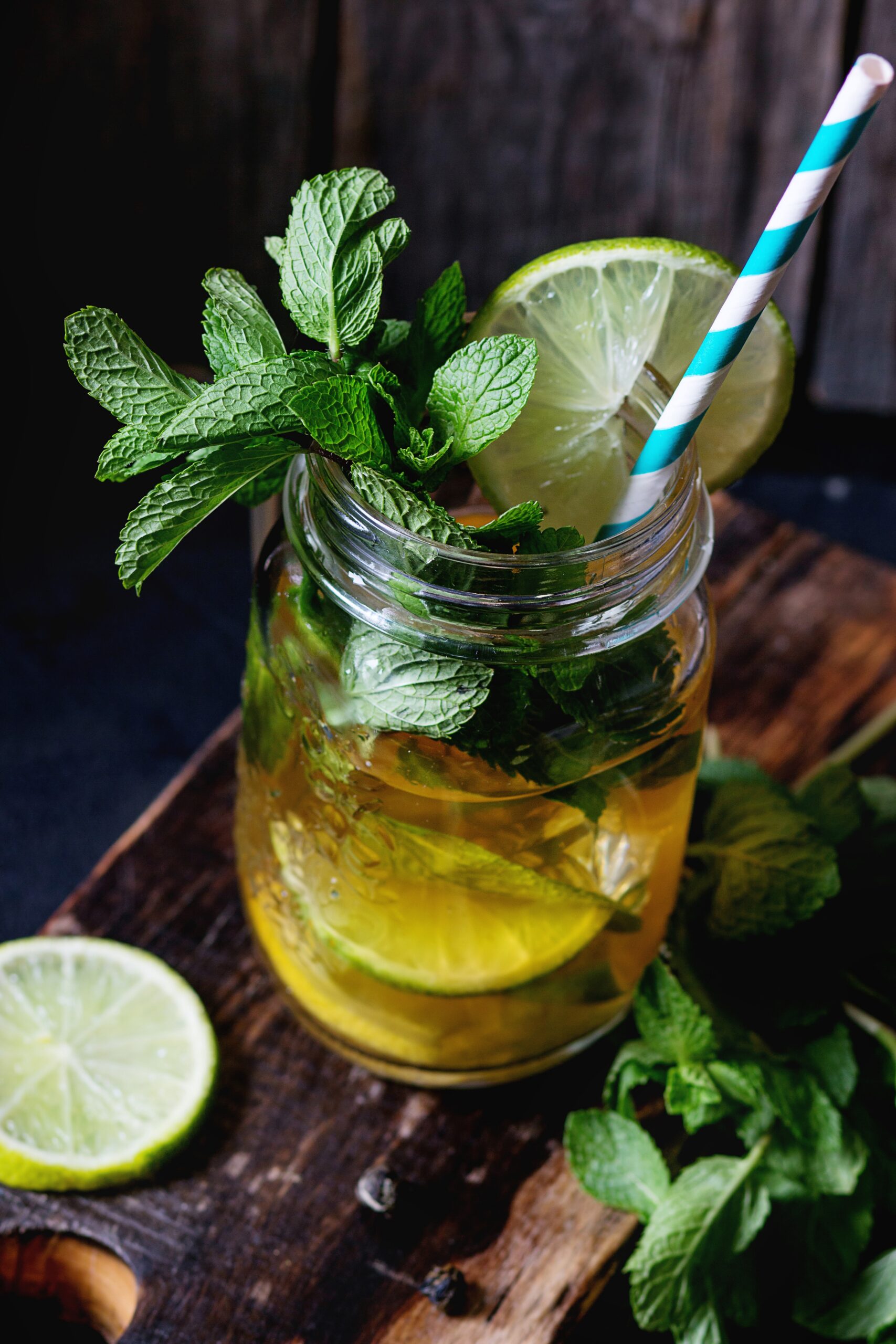 Ice green tea with ice, fresh mint, lime and lemon in glass mason jar with retro cocktail tube, served on wooden chopping board with old wooden background. Dark rustic style. Day light.