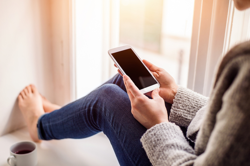 Unrecognizable woman sitting on window sill with smart phone