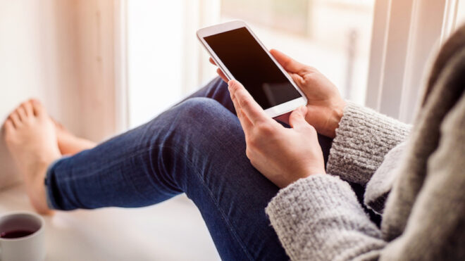 Unrecognizable woman sitting on window sill with smart phone