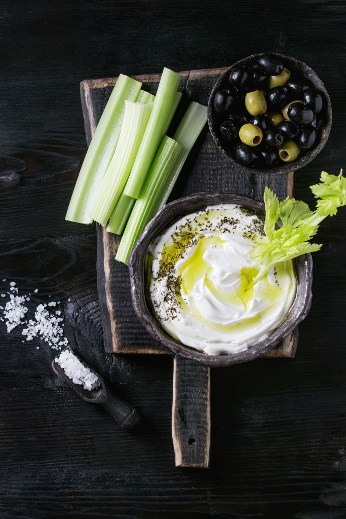 Fresh celery with yogurt and olive oil dip in ceramic bowl, served with sea salt and black, green olives on wood chopping board over black wooden burnt background. Top view with space. Healthy snack