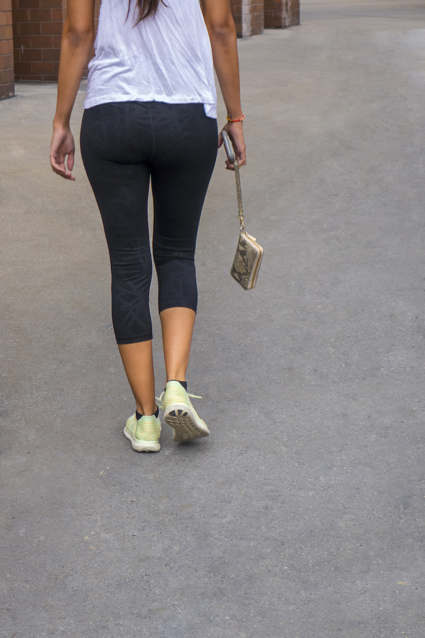 back of a woman walking holding a hanging pocketbook and cell phone in yoga pants sneakers on a city walkway