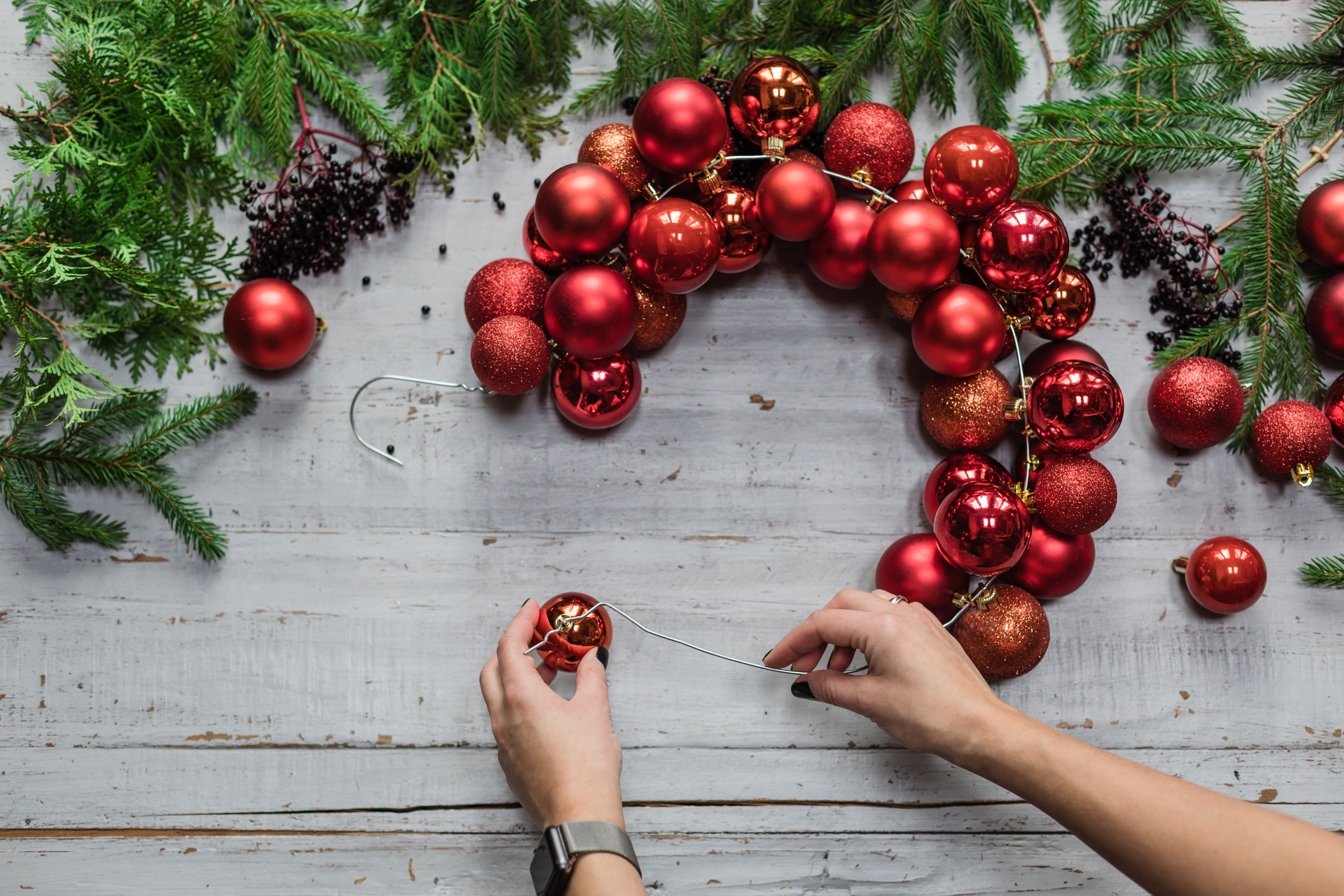 Top view of female hands making Christmas wreath with fir branches and decorative red balls on wooden rustic tabletop