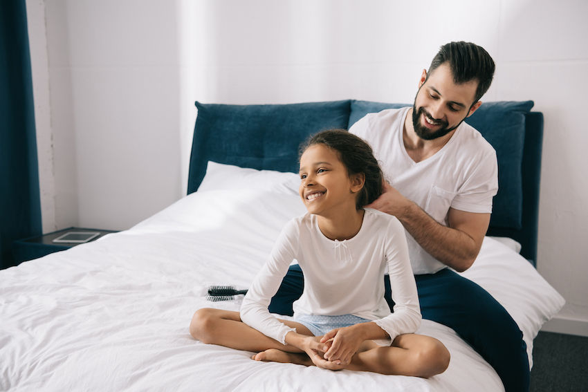 Happy bearded man combing hair of cute smiling daughter sitting on bed
