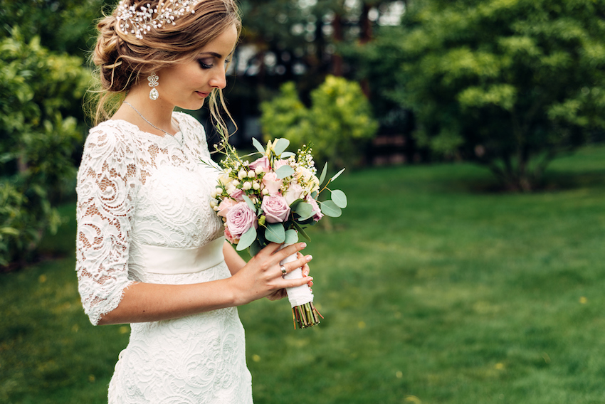 bride in a white long dress with a wedding bouquet is standing alone and looks at a bouquet waiting for her husband