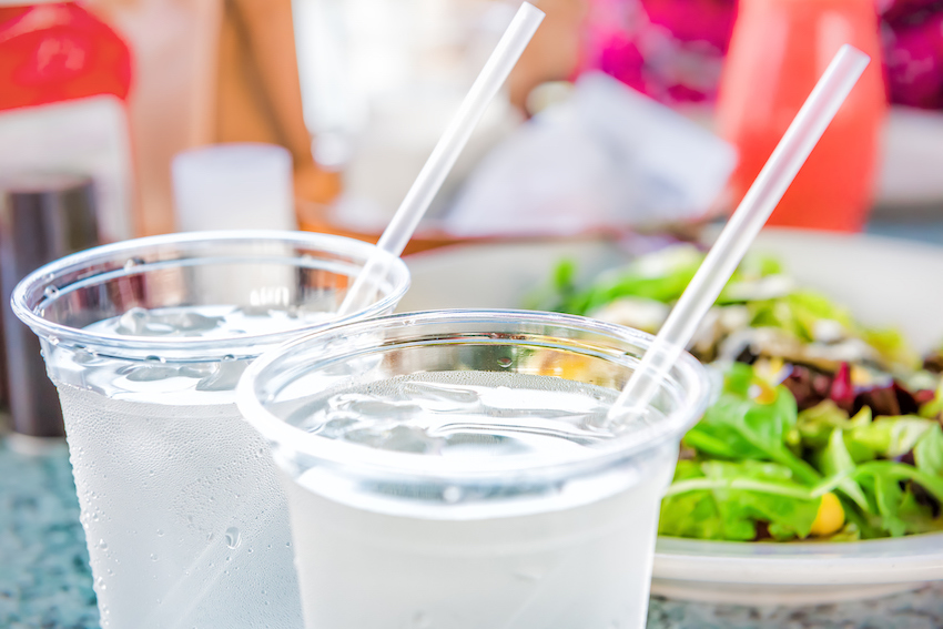 Macro closeup of two plastic cups with ice cubes and water and condensation on table with straws by green salad in restaurant