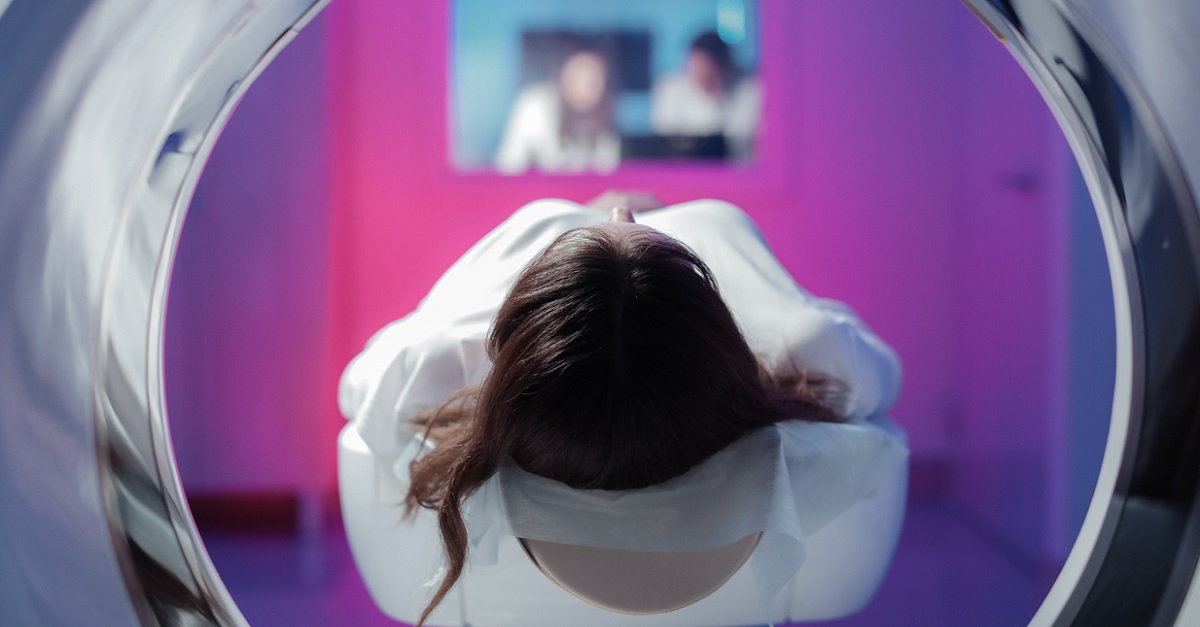 The girl patient is lying in the tomograph and waiting for a scan. Three doctors from the exam room look at the pictures