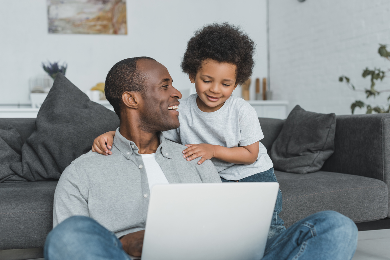 smiling african american father holding laptop and looking at son at home