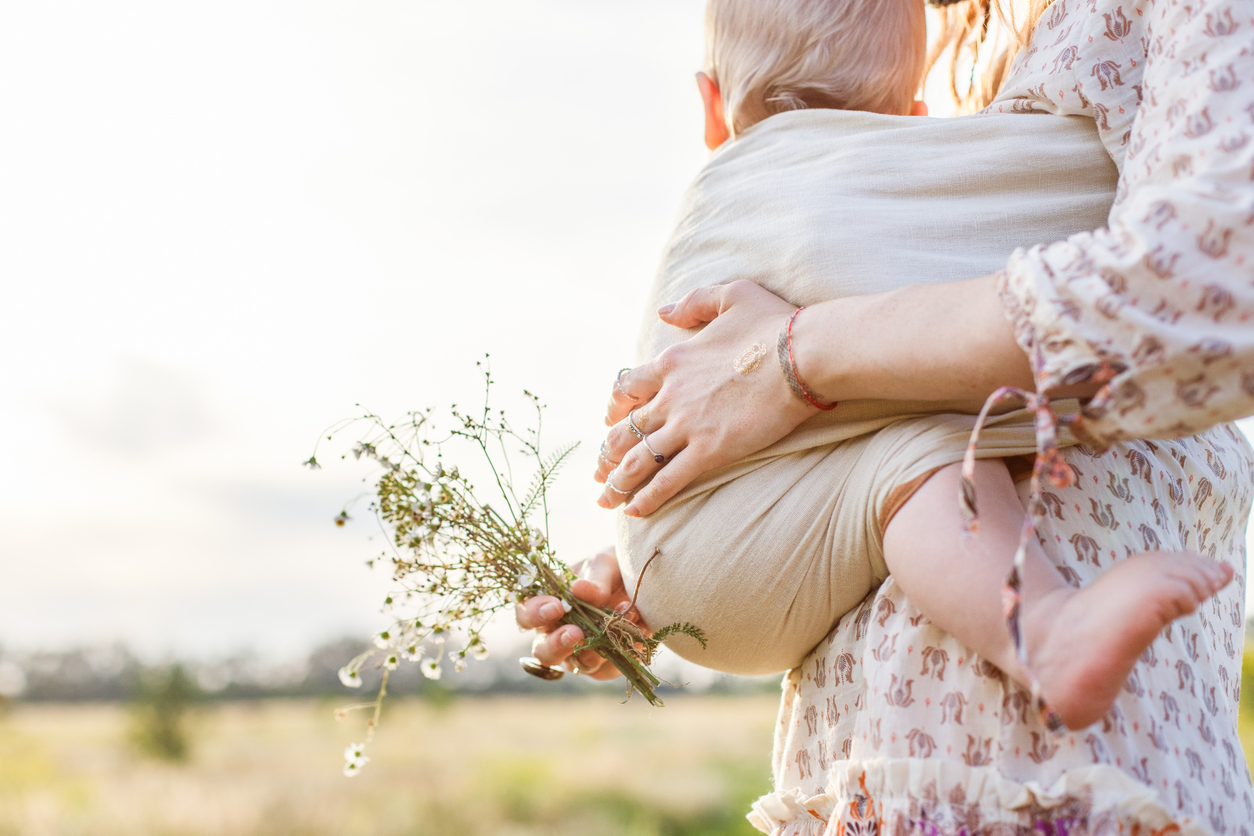 Little baby boy and his mother walking in the fields during summer day. Mother is holding and tickling her baby, babywearing in sling. Natural parenting concept