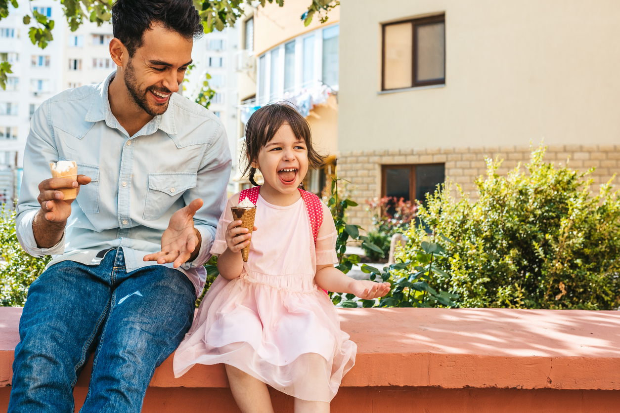 Image of happy cute little girl sitting with dad on the city street and eating ice-cream outdoor. Fun girl kid and father have fun and playing outside. Good relationship between dad and daughter