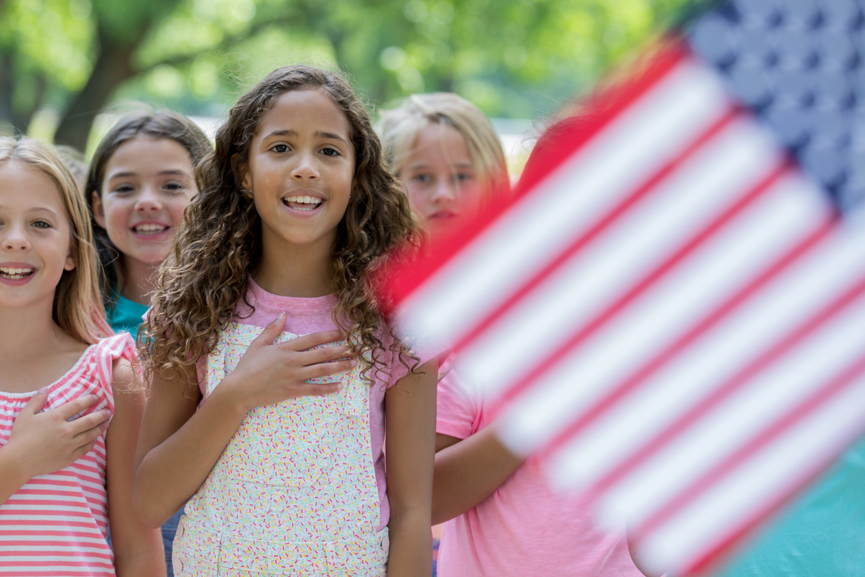 Biracial elementary age little girl reciting Pledge of Allegiance with classmates outdoors