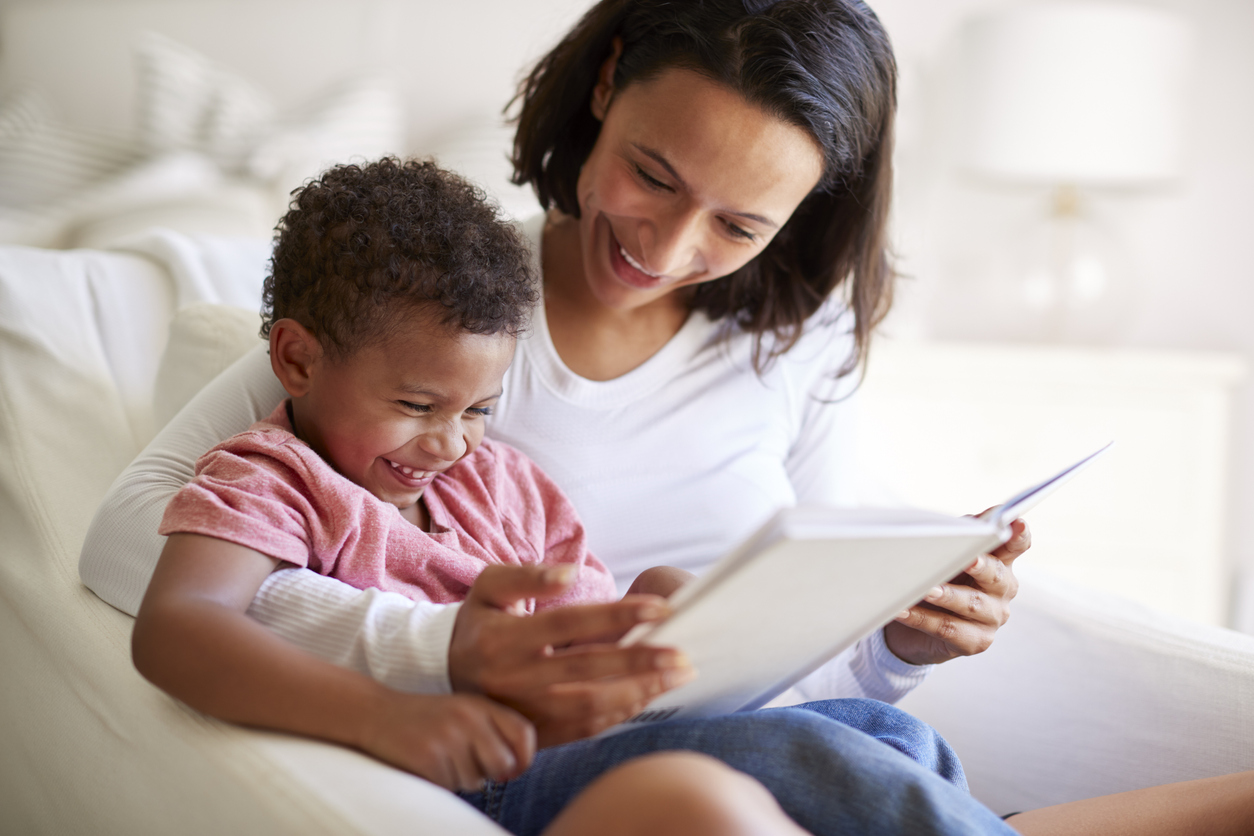 Close up of mixed race young adult mother sitting in an armchair reading a book with her three year old son on her knee, laughing, close up