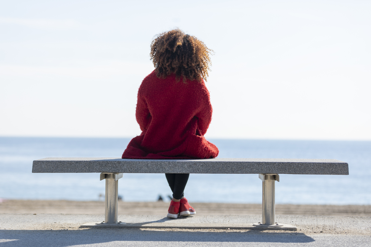 Rear view of a young curly woman wearing red denim jacket sitting on a bench while looking away to horizon over sea