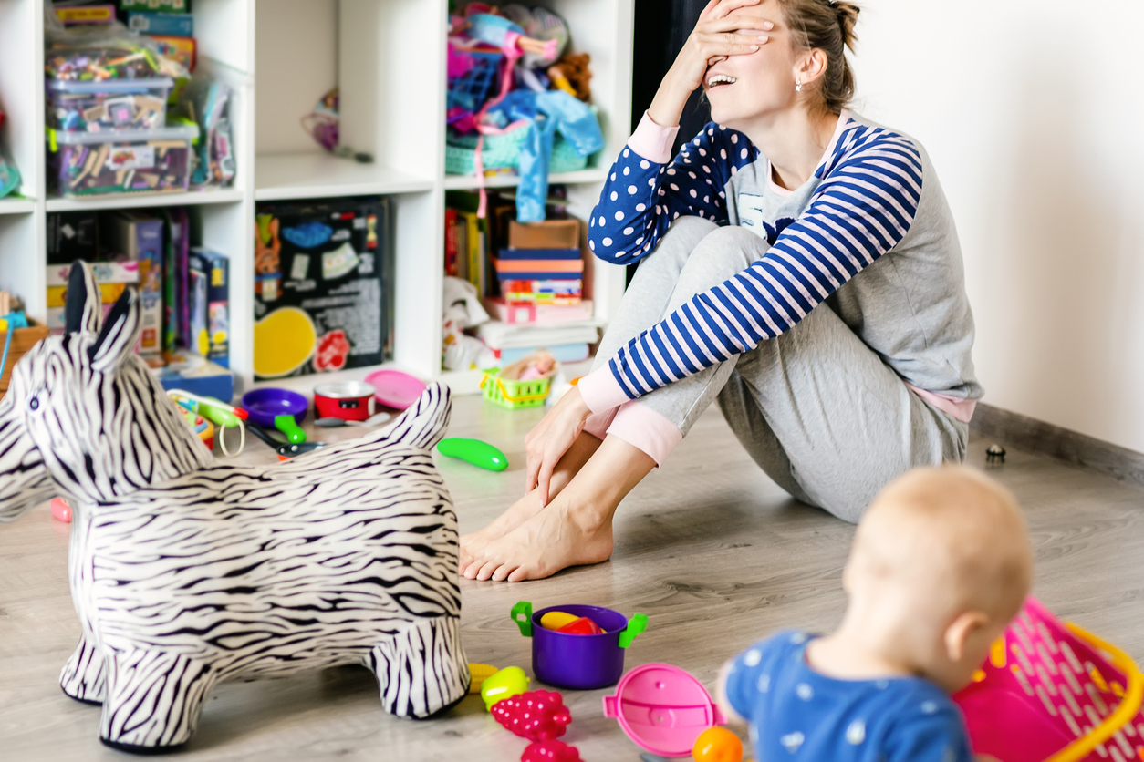 Tired of everyday household mother sitting on floor with hands on face. Kid playing in messy room. Scaterred toys and disorder. Happy parenting