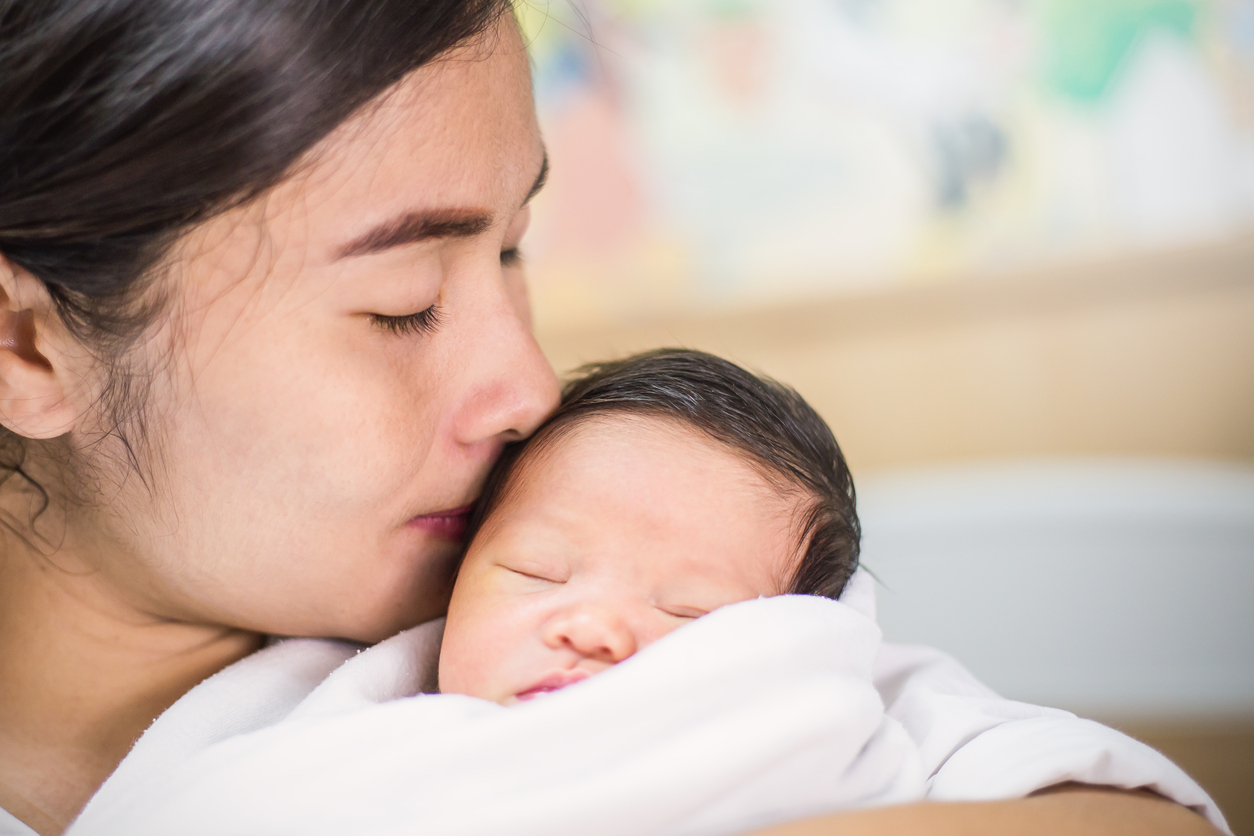 Portrait of beautiful young asian mother kissing  her newborn baby, copy space with bed in the hospital background.