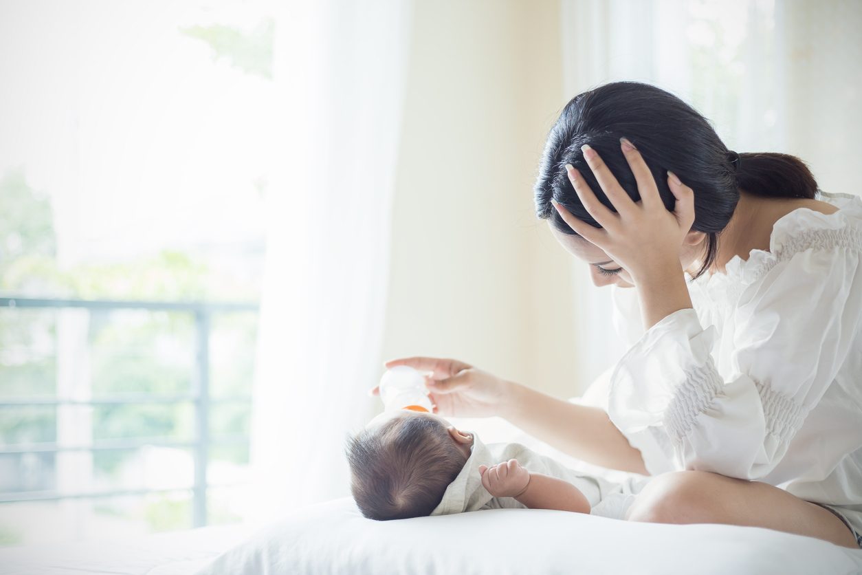 Asian Mother nursery feeding bottle of formula milk to newborn baby in bed suffering from post natal depression. Health care single mom motherhood stressful concept.