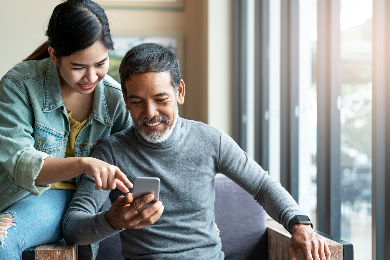 Attractive mature asian man with white stylish short beard looking at smartphone computer with teenage eye glasses hipster woman in cafe. Teaching internet online or wifi technology concept