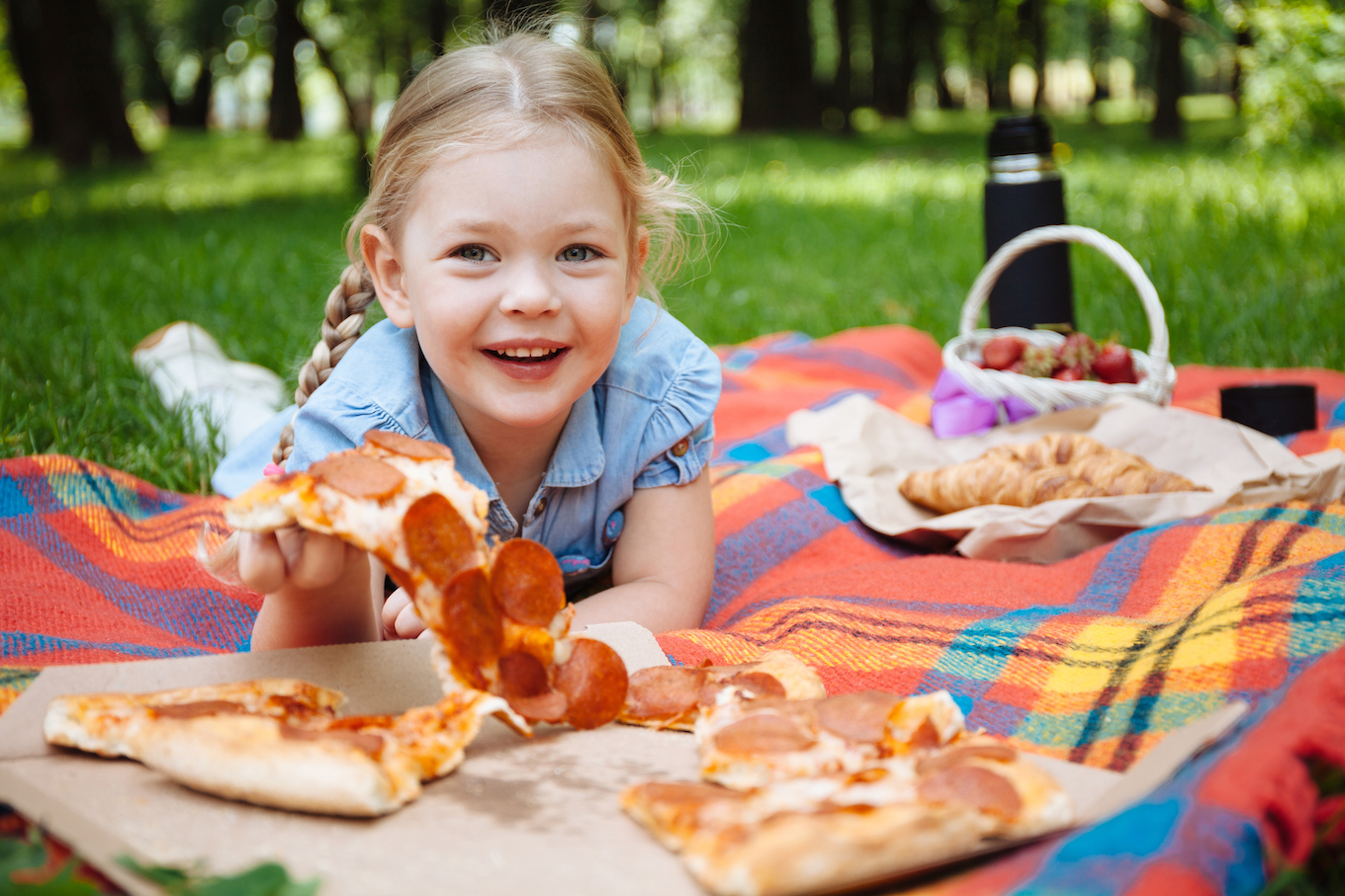 Little baby girl eating pizza and laughing outdoors on the background of green grass, summer picnic. Rest in the park