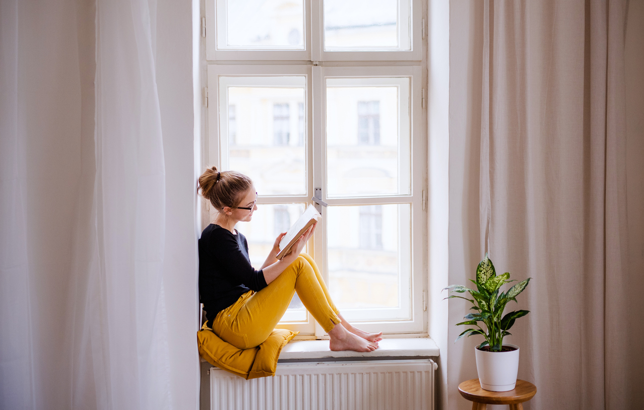 A young female student with a book sitting on window sill, studying.