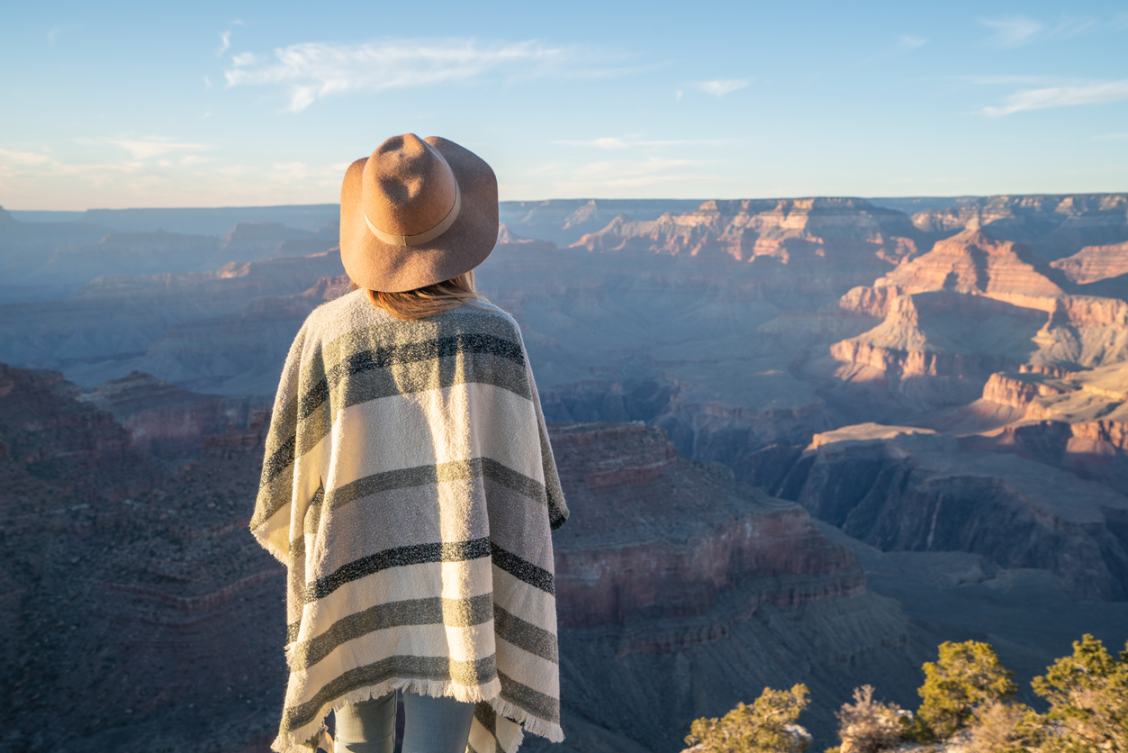 Rear view of unrecognisable woman standing above Grand Canyon on top of it all; girl with poncho and hat looking at landscape; fashionable internet people