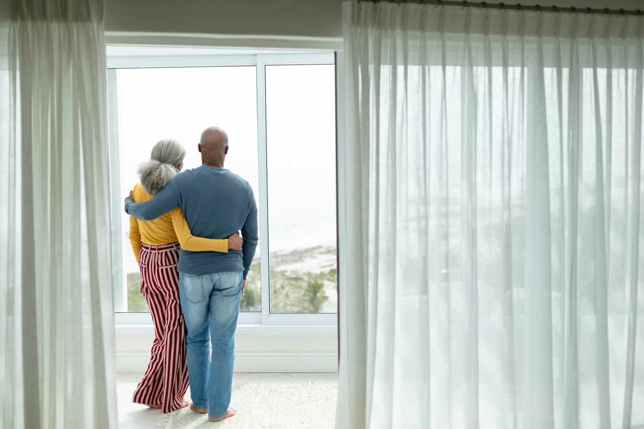 Active senior couple with arm around standing near window at home