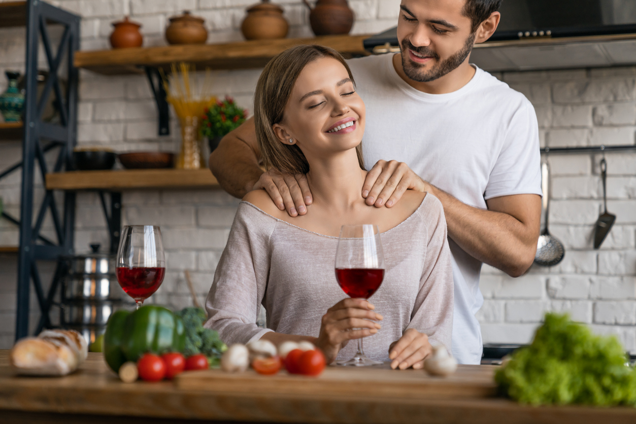 Beautiful girl drinking wine while handsome man making massage to her in home kitchen