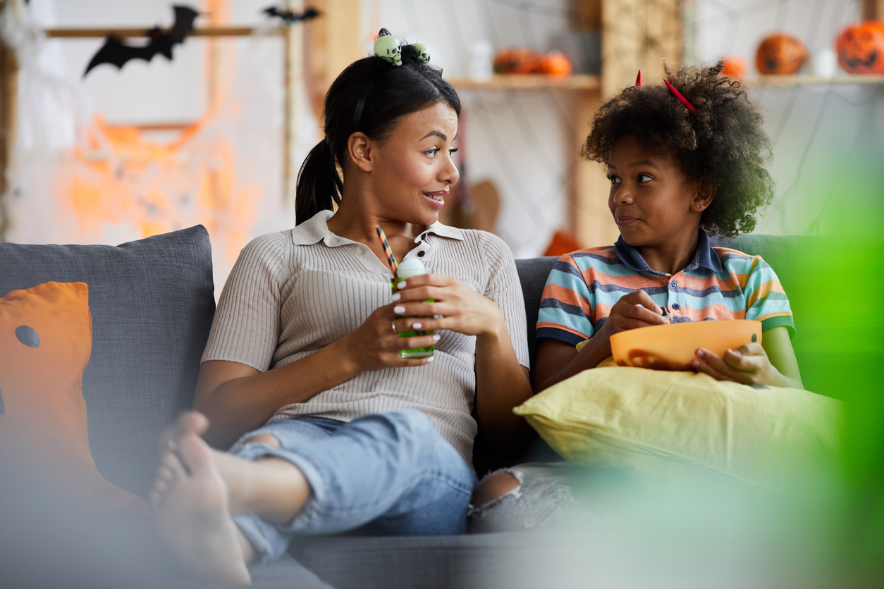 Positive Afro-American mother and son sitting on sofa and chatting while drinking cocktail and eating candies on Halloween