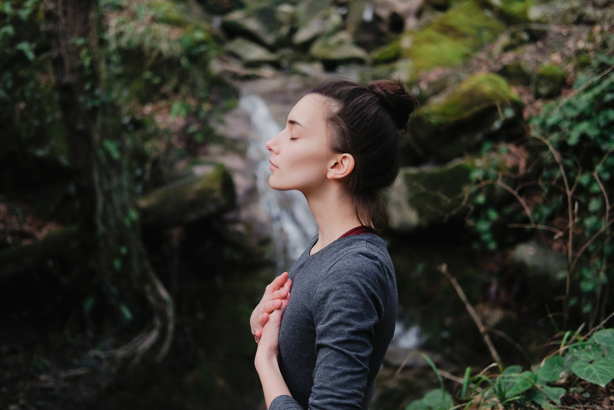 Young woman practicing breathing yoga pranayama outdoors in moss forest on background of waterfall. Unity with nature concept.