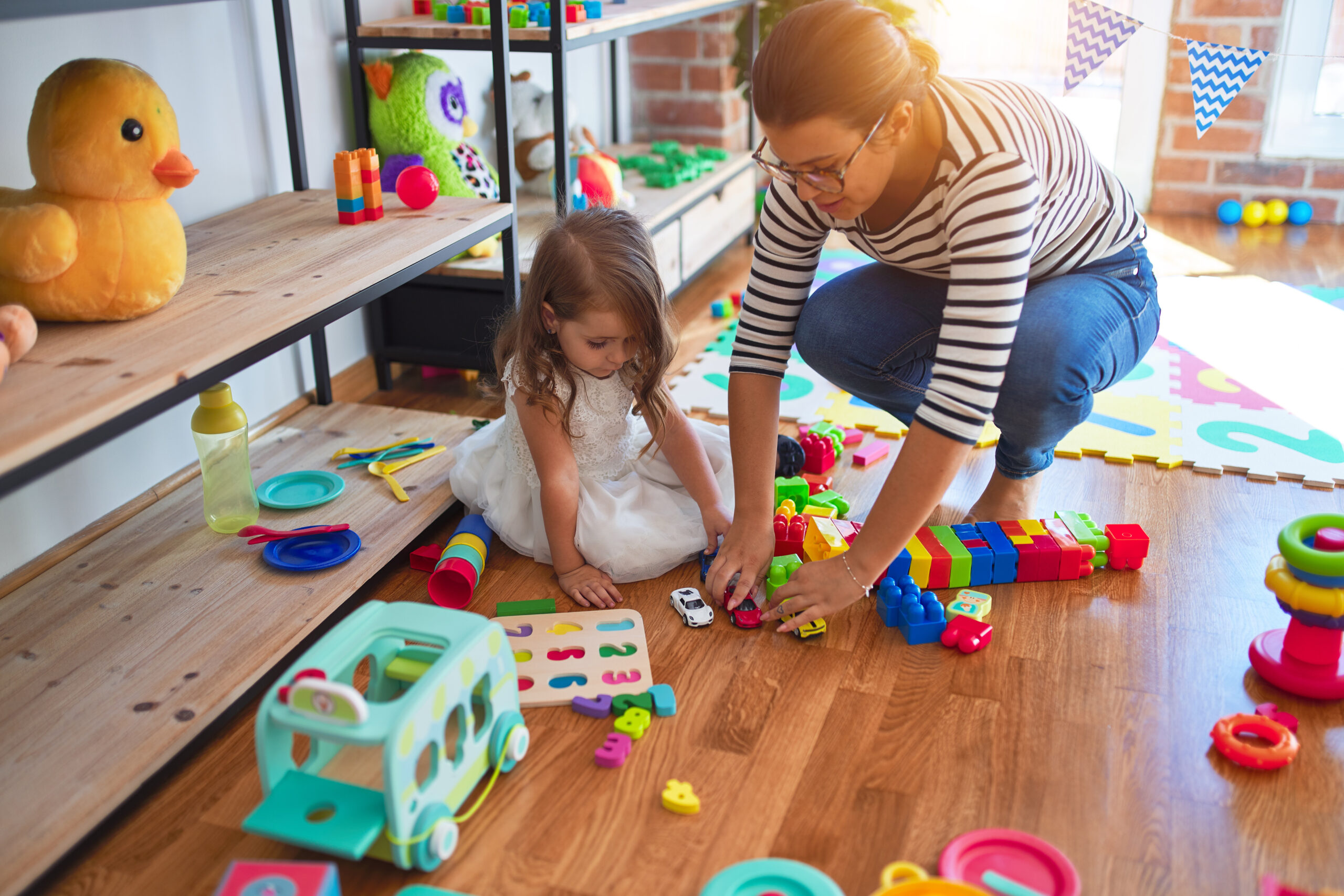 Beautiful teacher woman and toddler playing around lots of toys at kindergarten