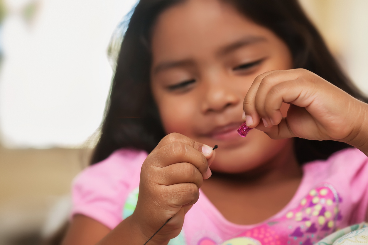 A cute little girl having fun while while creating bead jewelry using string and colorful beads.