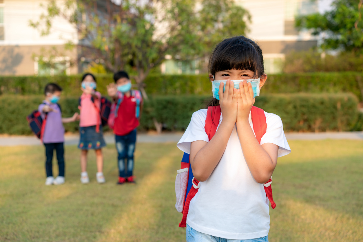 Asian girl preschool child student wearing healthy face mask sneeze with friends in background. Social distancing recommends at least one meter of distance between yourself and people who are coughing or sneezing.