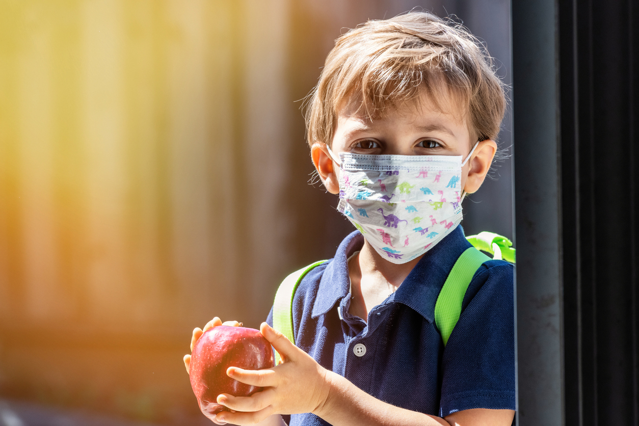 Little schoolboy holding an apple wearing a protective mask and a backpack