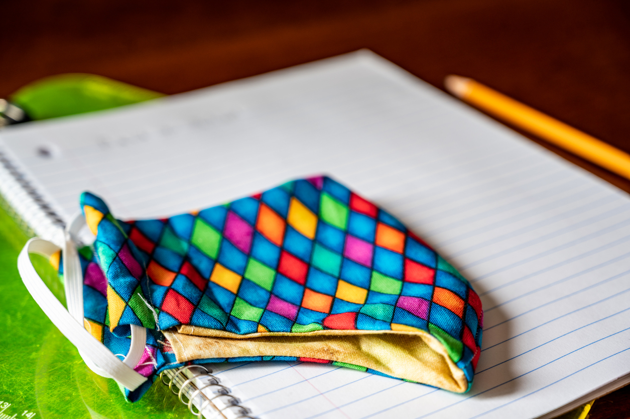 Face mask resting on top of back to school supplies of paper, pencil and a clip board